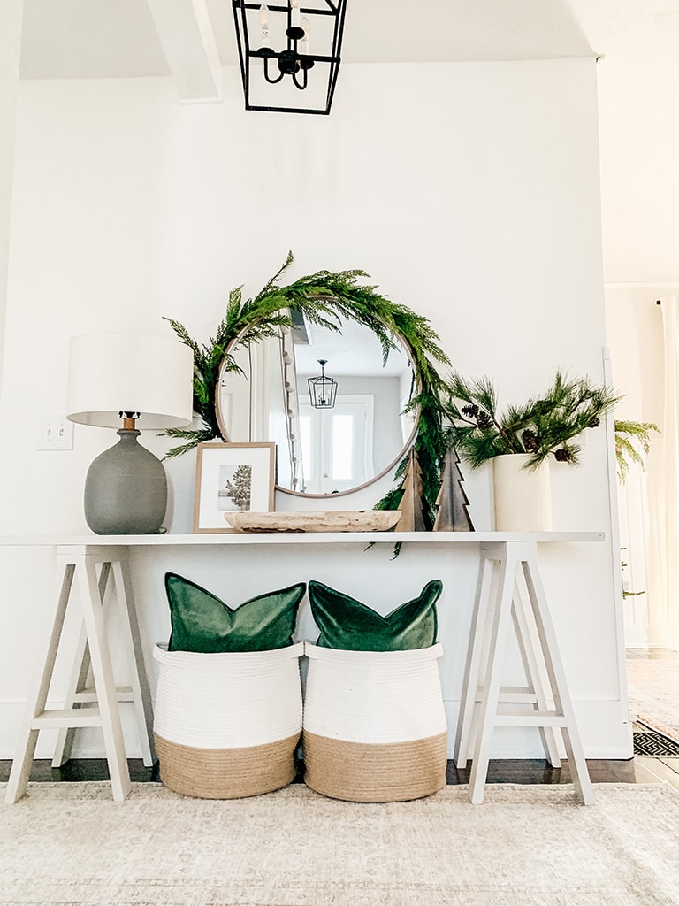 Grey Console table decorated for Christmas Featuring garland wrapped around a round mirror, green velvet pillows, metal chrsitmas trees and pine stems in a cream vase.