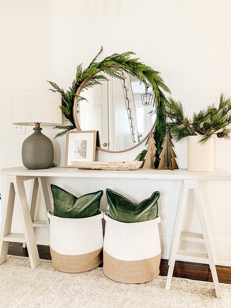 Grey Console table decorated for Christmas Featuring garland wrapped around a round mirror, green velvet pillows, metal chrsitmas trees and pine stems in a cream vase.