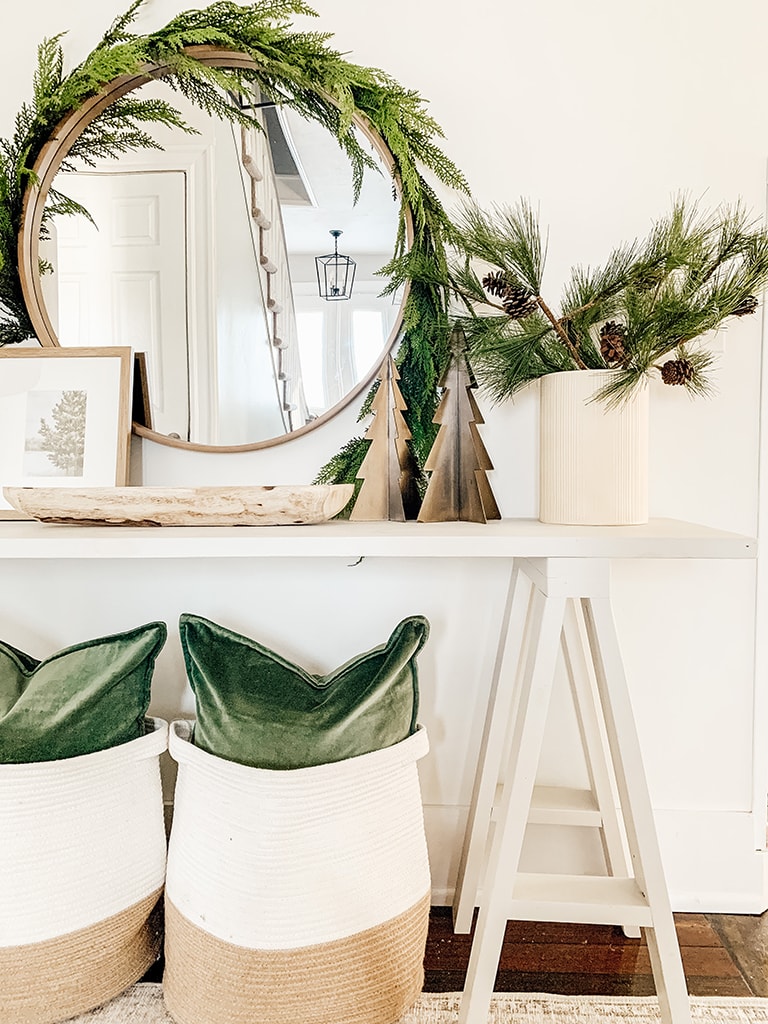 Grey Console table decorated for Christmas Featuring garland wrapped around a round mirror, green velvet pillows, metal chrsitmas trees and pine stems in a cream vase.