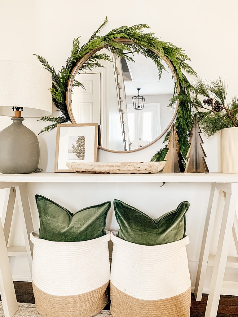 Grey Console table decorated for Christmas Featuring garland wrapped around a round mirror, green velvet pillows, metal chrsitmas trees and pine stems in a cream vase.