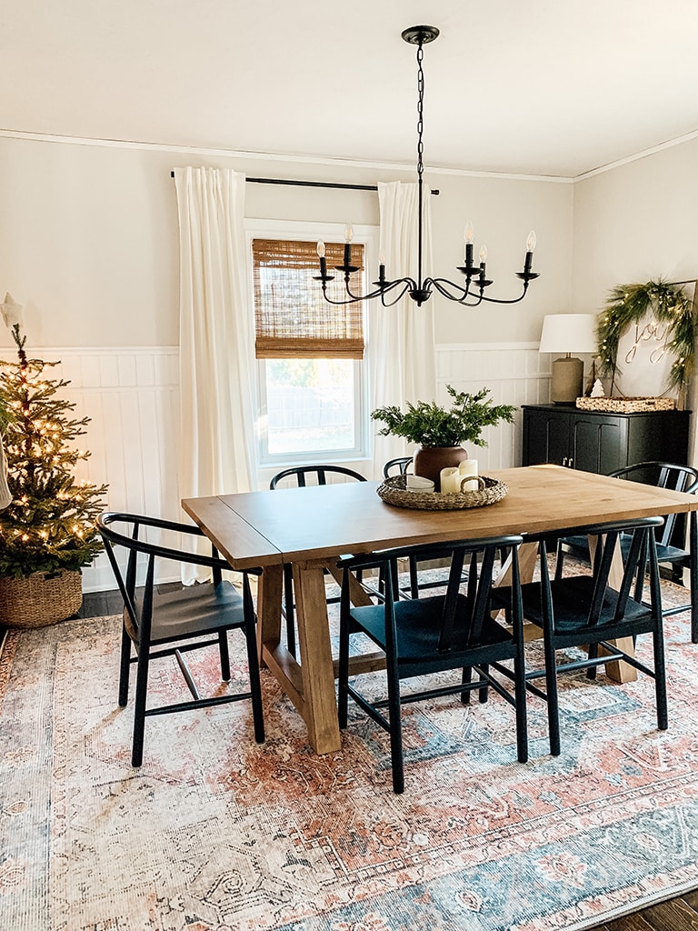 Christmas Dining Room featuring a small lit christmas tree, large black chandelier, wreath lit up with the words joy, wood table and black dining chairs.