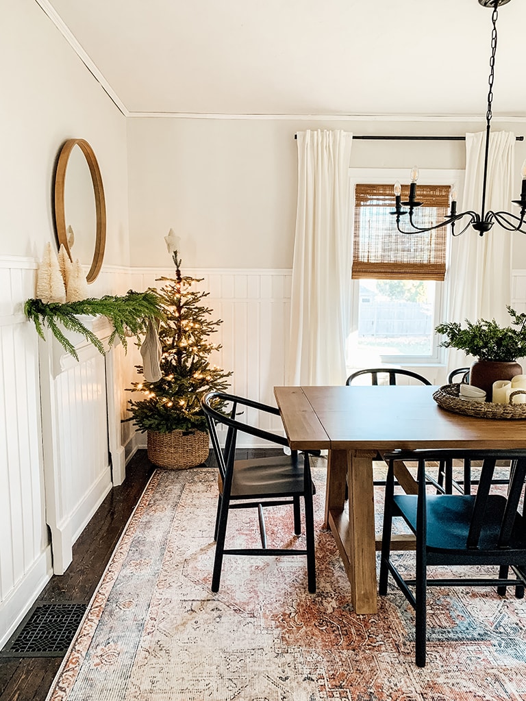 Christmas Dining Room featuring a small lit christmas tree, large black chandelier, wreath lit up with the words joy, wood table and black dining chairs.