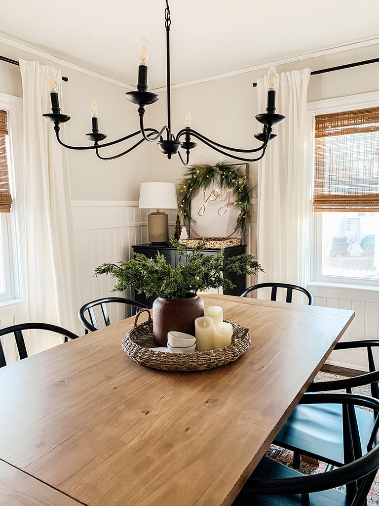 Christmas Dining Room featuring a small lit christmas tree, large black chandelier, wreath lit up with the words joy, wood table and black dining chairs.