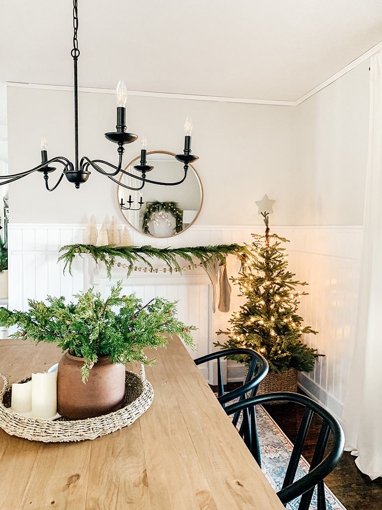 Christmas Dining Room featuring a small lit christmas tree, large black chandelier, wreath lit up with the words joy, wood table and black dining chairs.