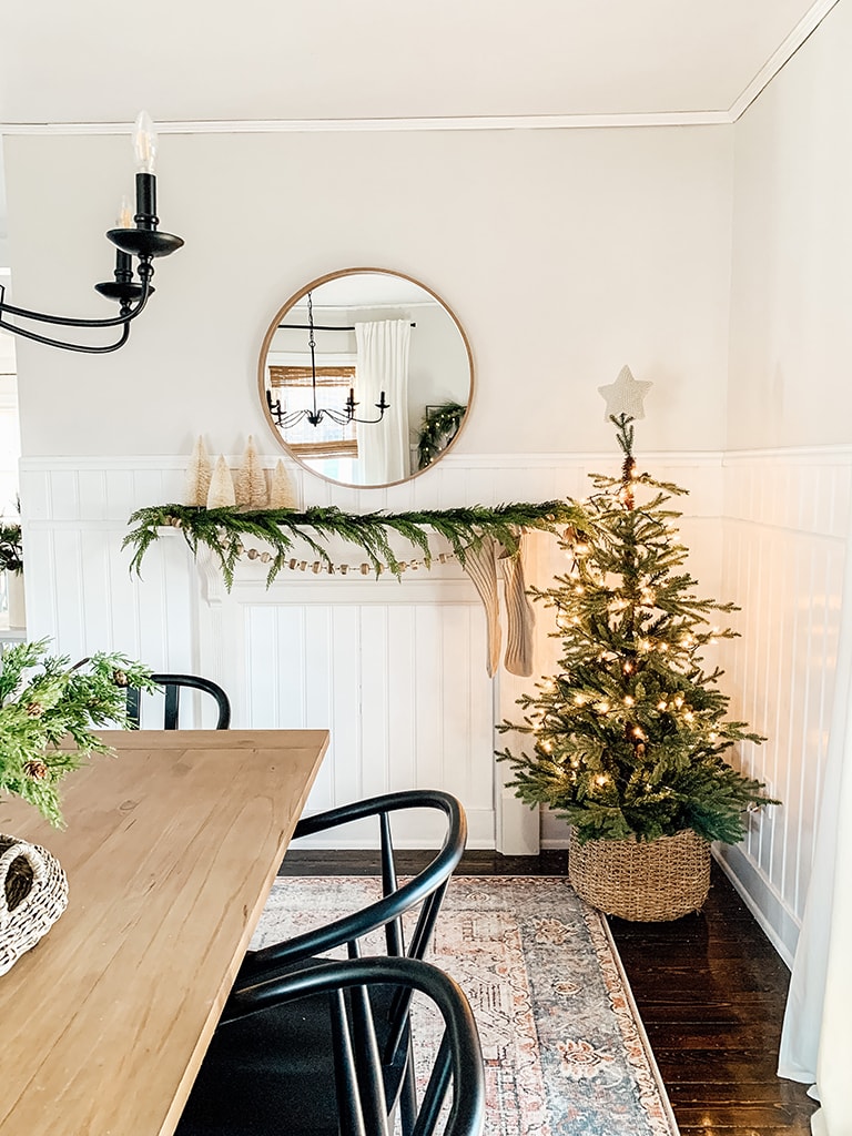 Christmas Dining Room featuring a small lit christmas tree, large black chandelier, wreath lit up with the words joy, wood table and black dining chairs.