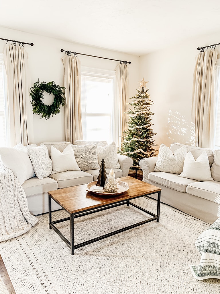 Simple neutral Christmas living room with slip cover couches a lit artificial tree in the corner and a cedar wreath on the wall.