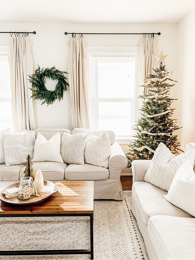 Simple neutral Christmas living room with slip cover couches a lit artificial tree in the corner and a cedar wreath on the wall.